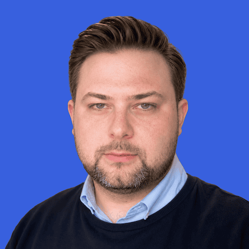 A man with short brown hair and beard wearing a light blue collared shirt and dark sweater against a solid blue background.