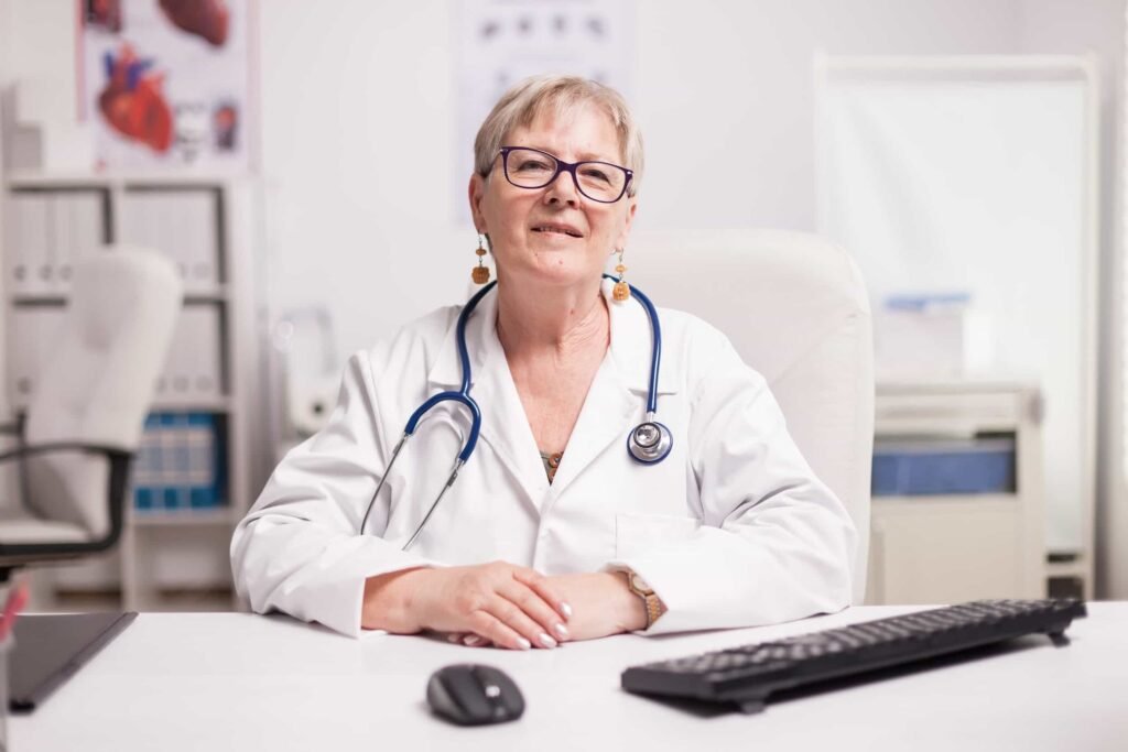 An older woman doctor offering Medical Services, wearing a white coat and stethoscope, sits at a desk with a keyboard and mouse in a medical office. Medical SEO