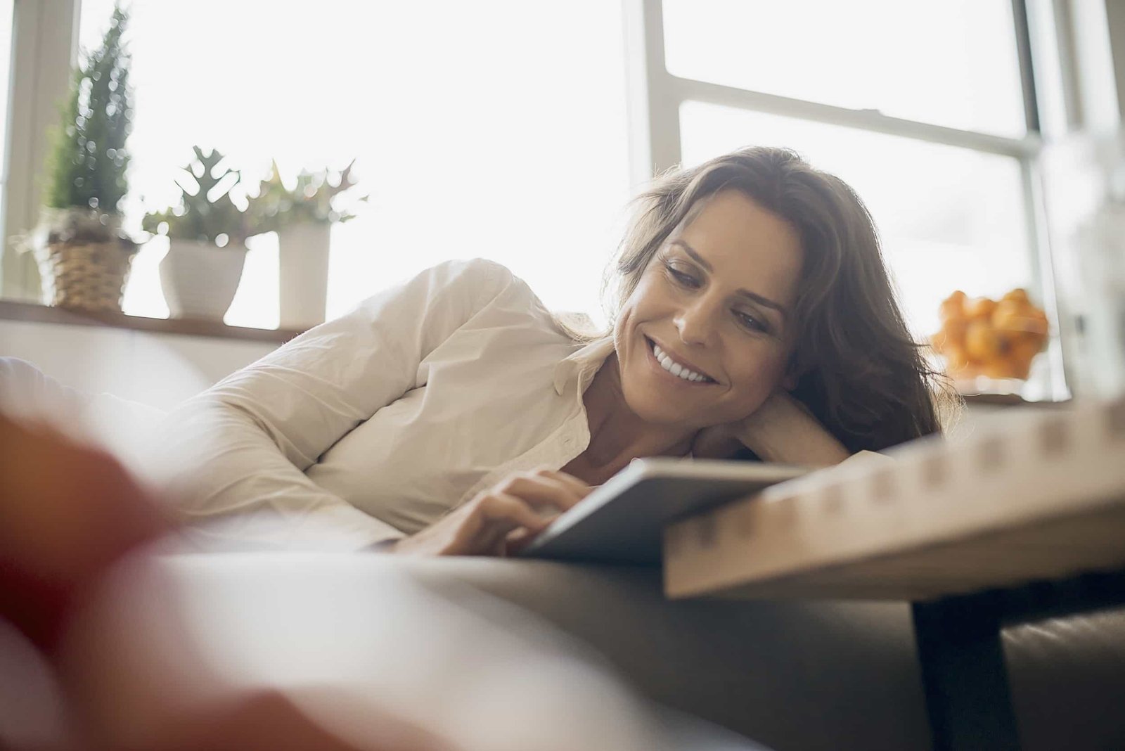 Woman lying on a couch, smiling and reading a tablet about conversion rate optimization. Potted plants and a window create a relaxed backdrop, perfect for exploring new CRO strategies. Queens NY