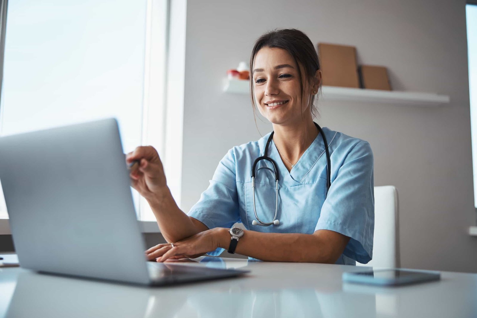 A healthcare professional in scrubs with a stethoscope sits at a desk, smiling and pointing at a laptop screen in a well-lit room. Is SEO the Same as GEO? Here's What You Need to Know