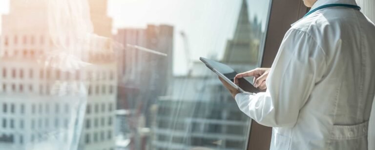 A person in a white lab coat uses a tablet while standing near a window overlooking city buildings. When Your Medical Practice Website Is Costing You