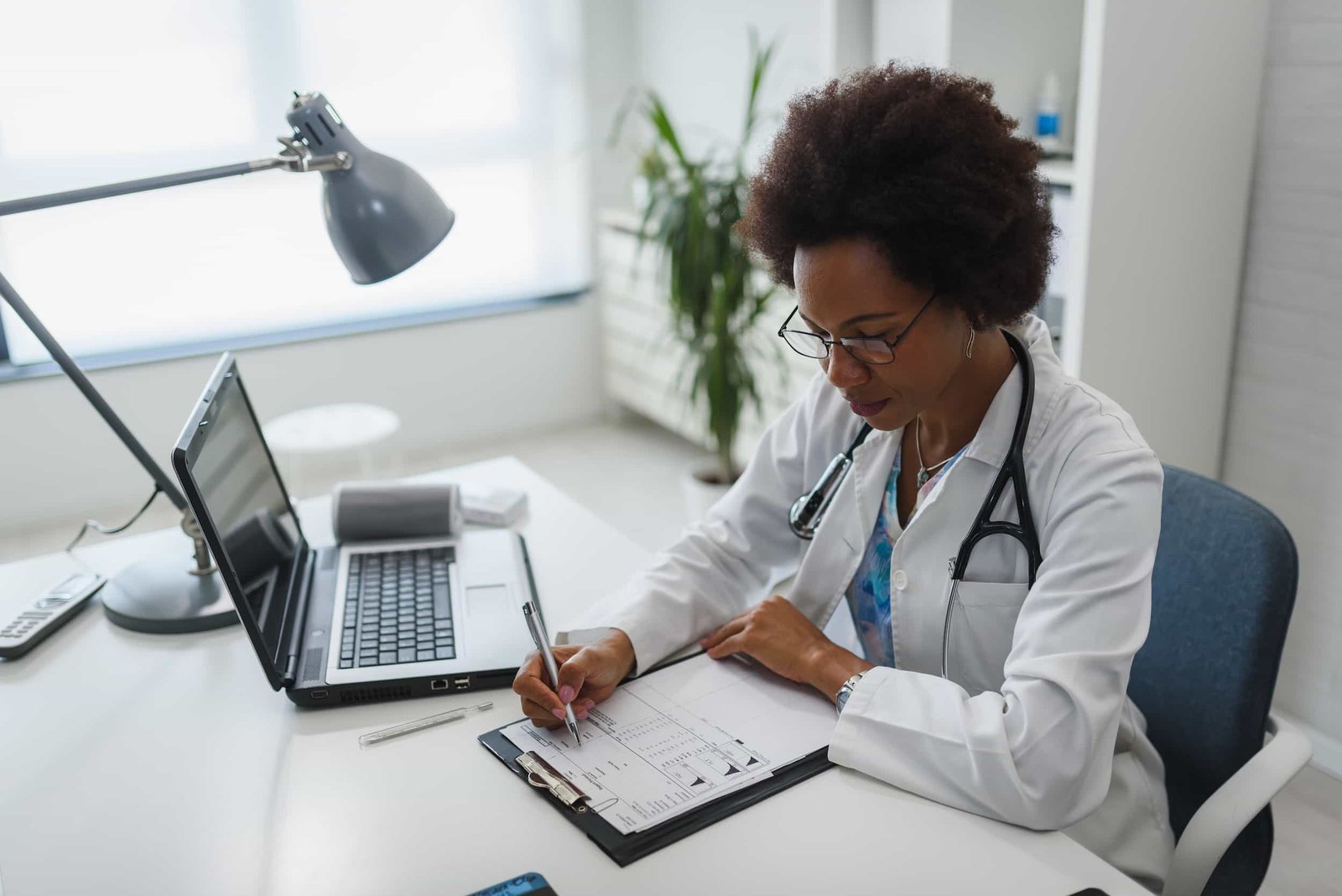 A doctor wearing a white coat and stethoscope fills out paperwork at a desk with a laptop, lamp, and clipboard in a medical office.