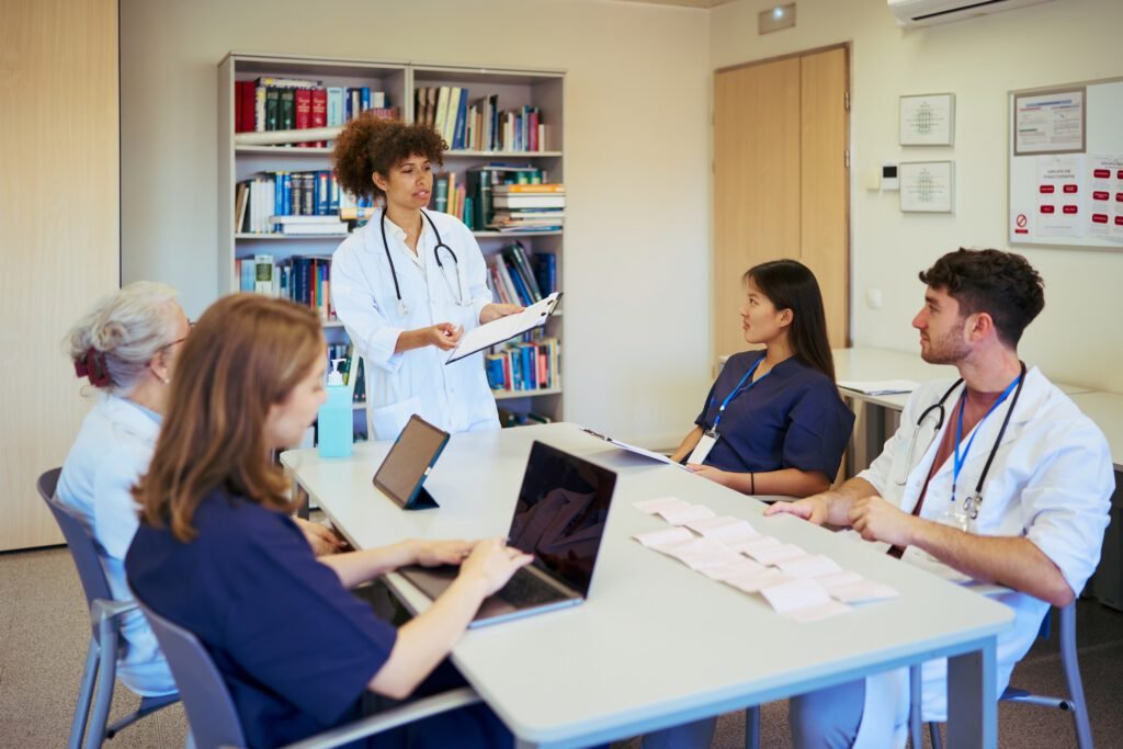 A doctor stands and presents to four colleagues seated at a table in a medical office, with laptops, papers, and books visible—demonstrating the collaborative spirit that even a Medical Web Designer in NYC might value for effective SEO strategies in healthcare.