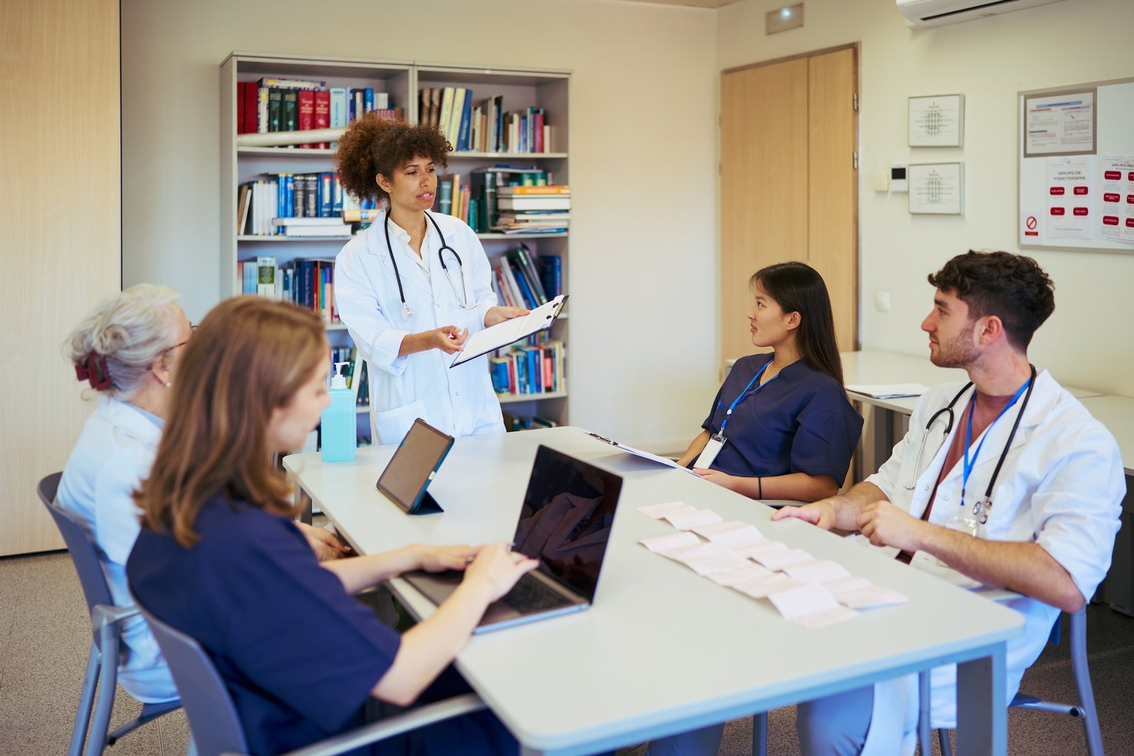 A doctor stands and presents to four colleagues seated at a table in a medical office, with laptops, papers, and books visible—demonstrating the collaborative spirit that even a Medical Web Designer in NYC might value for effective SEO strategies in healthcare.