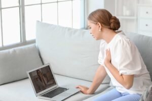 A woman sits on a couch having a video call on her laptop, with another woman visible on the laptop screen. Promise.