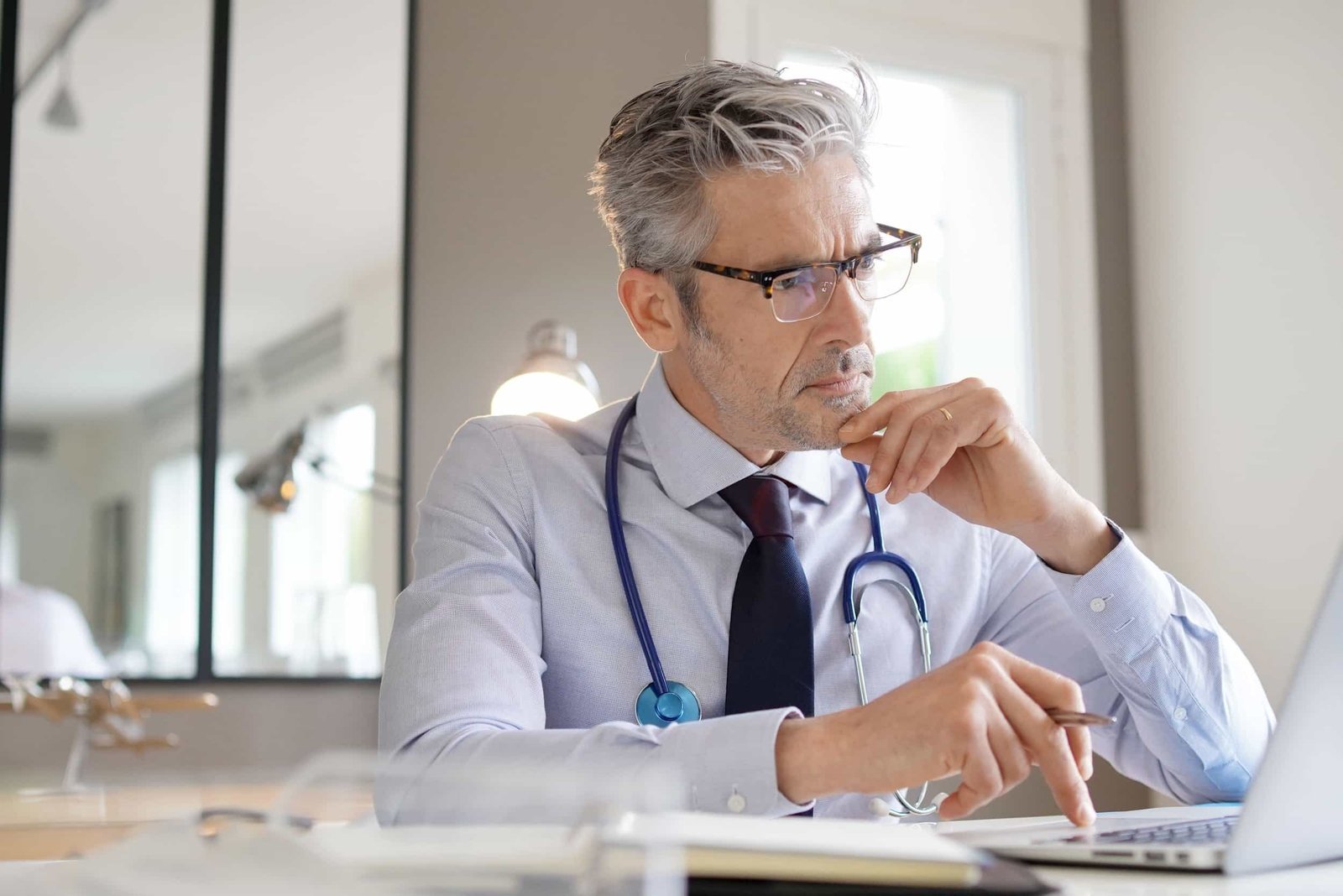 A doctor with gray hair and glasses, wearing a stethoscope and business attire, sits at a desk looking thoughtfully at a laptop screen.