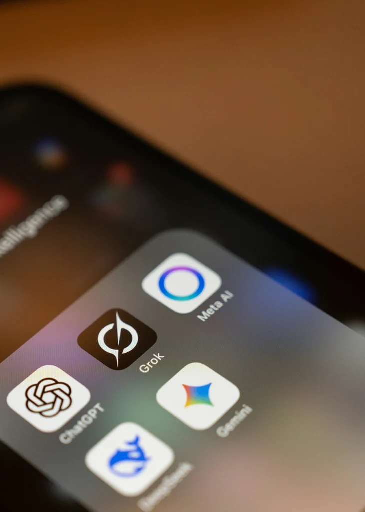 Close-up of a smartphone screen displaying the app icons for ChatGPT, Grok, Meta AI, and Gemini in a folder. AI Optimization (GEO/AEO)