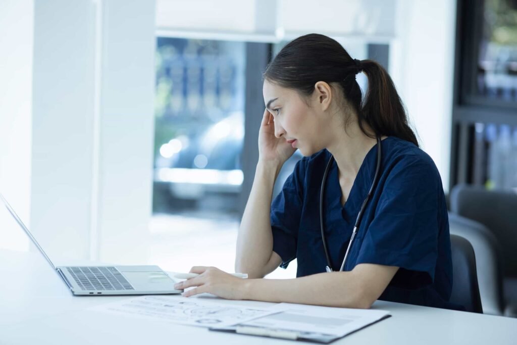 A woman in medical scrubs sits at a desk, looking at a laptop with a concerned expression, resting her head on one hand. Medical papers and a stethoscope are on the desk, suggesting she might be consulting with a website designer about creating an online platform for her practice. Before You Hire a Website Designer, Read This.