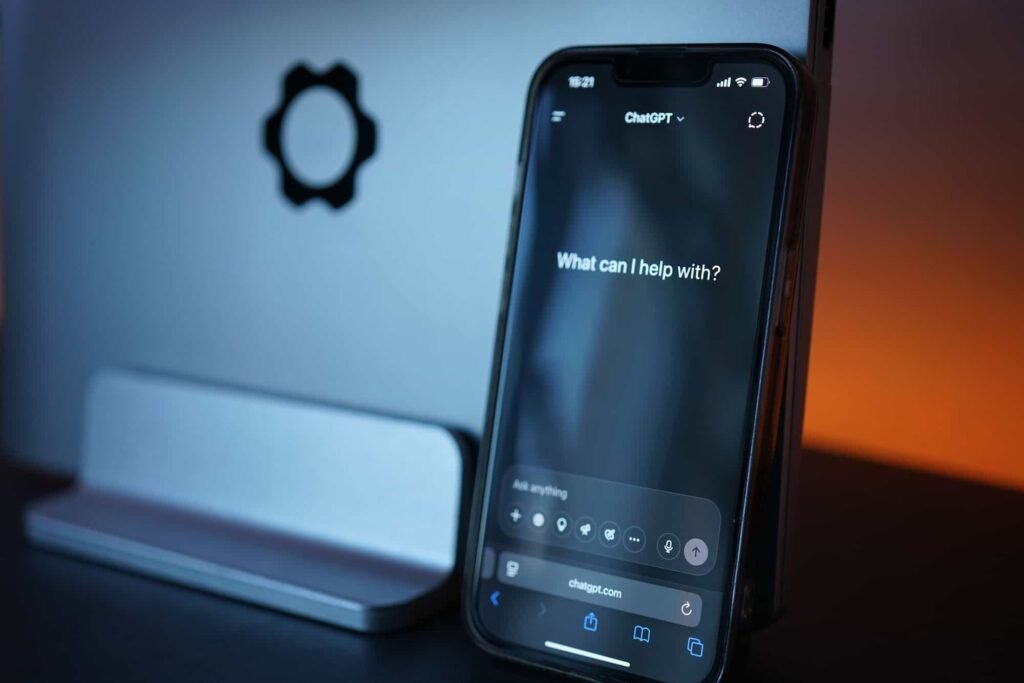 A smartphone displaying a ChatGPT interface that reads "What can I help with?" is propped up against a silver laptop with a gear symbol. Medical AI