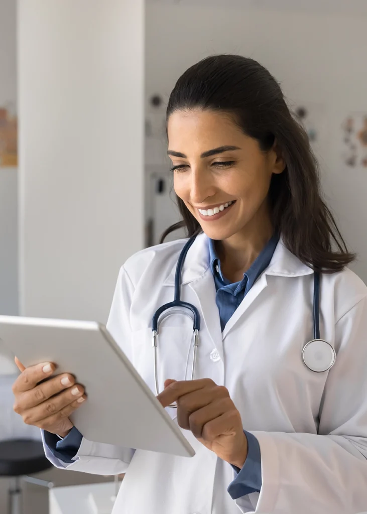 A doctor wearing a white coat and stethoscope smiles while using a digital tablet in a bright medical office. Conversion Rate Optimization in New York City. CRO
