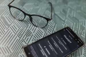 A pair of eyeglasses and a smartphone displaying the ChatGPT interface rest on a patterned surface, highlighting the growing connection between AI technology and medical innovation. AI Medical in Queens
