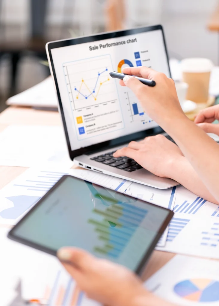 Two people review sales performance charts on a laptop while one person holds a tablet displaying a bar graph.
