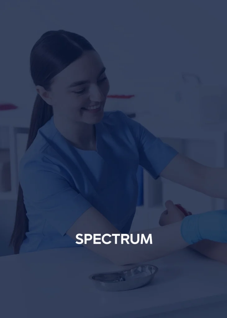 A healthcare worker in blue scrubs smiles while assisting a patient in a medical setting, embodying the compassionate care found throughout NYC. The word "SPECTRUM" appears over the image.