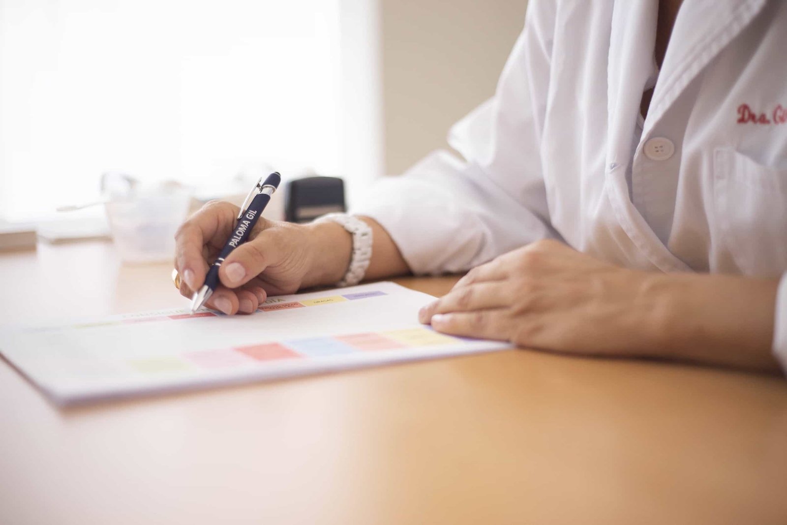 Person in a white lab coat writes on a color-coded document at a desk, with only their hands and torso visible. Healthcare Marketing Strategies That Fill Your Appointment Book.