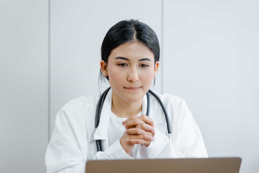 A person wearing a white coat and stethoscope sits at a desk with hands clasped, looking at a laptop. Discover proven patient acquisition strategies to grow your medical practice. Attract more high-value patients with tactics that actually work in today's digital landscape