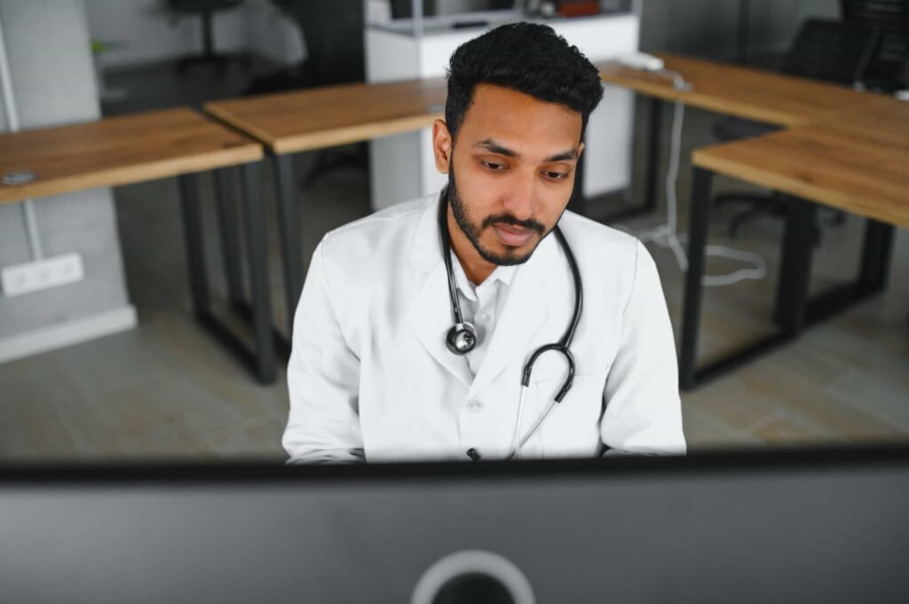 A doctor with a stethoscope around his neck sits at a desk, looking at a computer screen in a modern office setting. Google Business Profile for Medical Practices: Get Patients