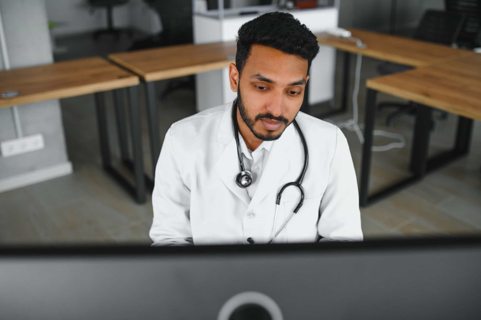A doctor with a stethoscope around his neck sits at a desk, looking at a computer screen in a modern office setting. Google Business Profile for Medical Practices: Get Patients
