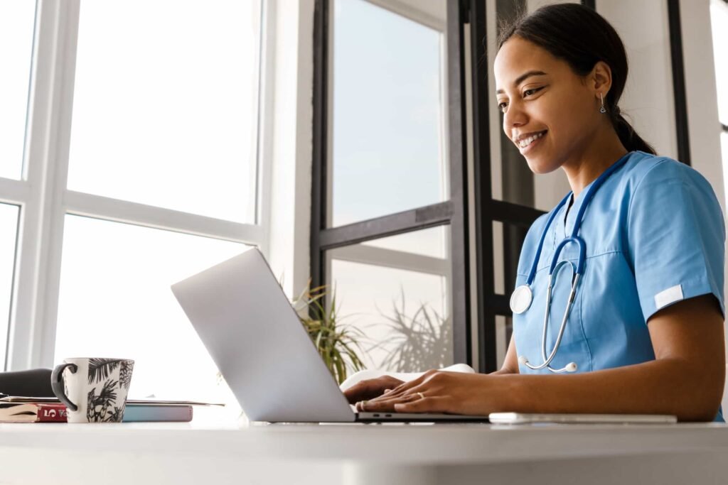 A smiling healthcare professional in blue scrubs with a stethoscope around their neck works on a laptop at a desk.