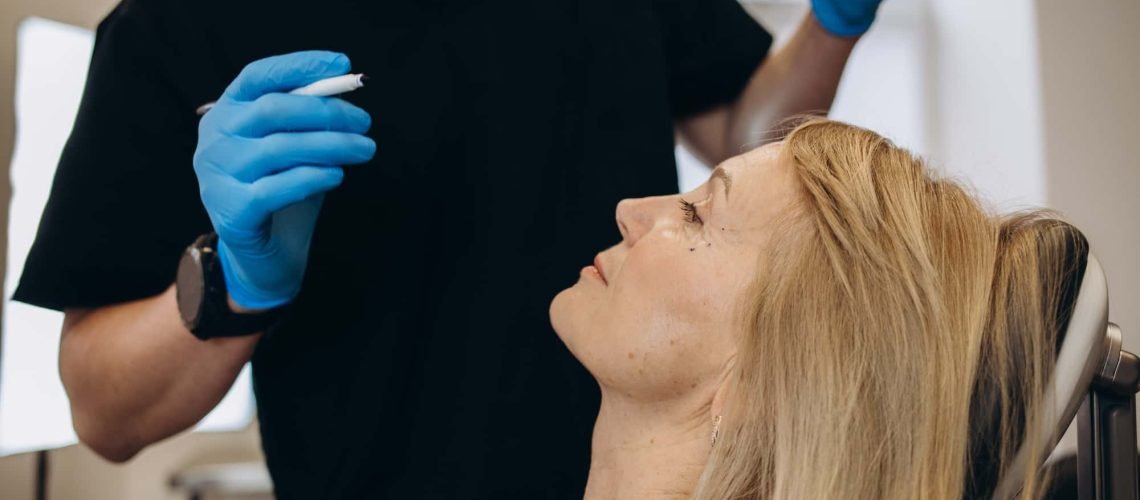 A woman sits in a chair looking up at a person wearing a black shirt and blue gloves, who is holding a small object near her face.