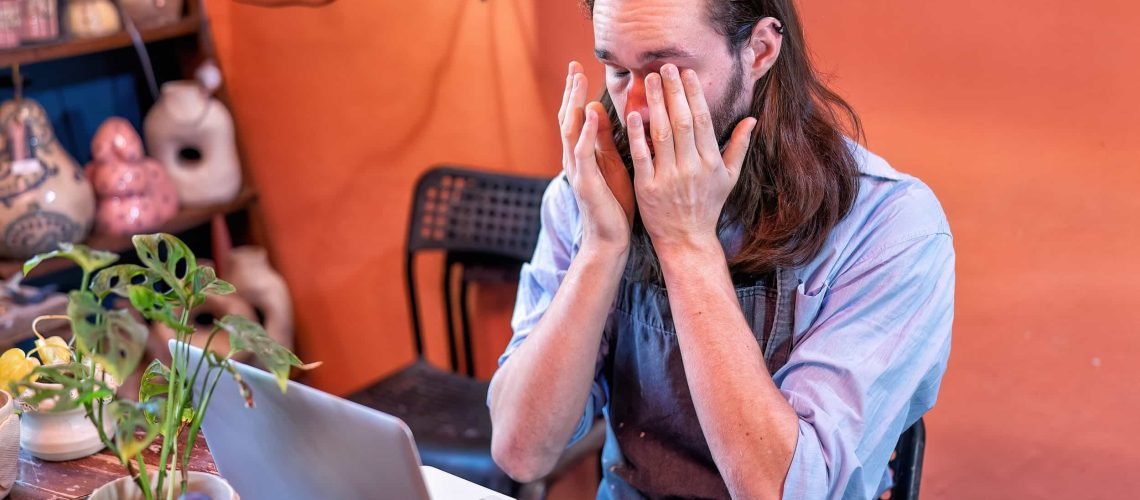 A person wearing an apron sits at a desk with a laptop, rubbing their eyes with both hands, appearing tired or stressed.