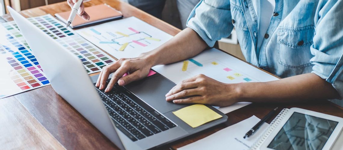 Two people collaborate at a desk with a laptop, color swatches, printed charts, a notebook, and a tablet, discussing design or project plans.