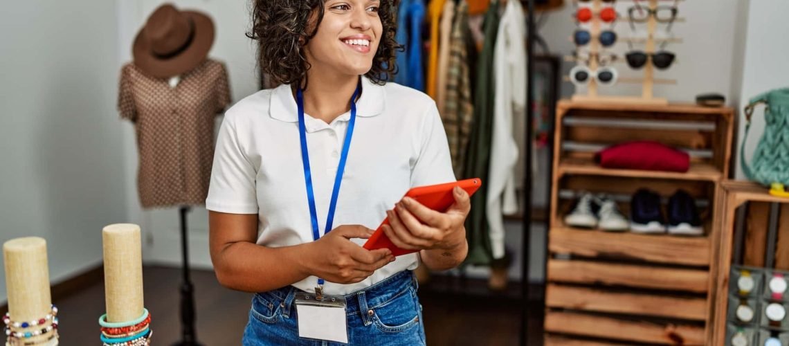 A person with curly hair and a lanyard holds a tablet in a clothing store. Displayed are clothes, shoes, accessories, and hats.