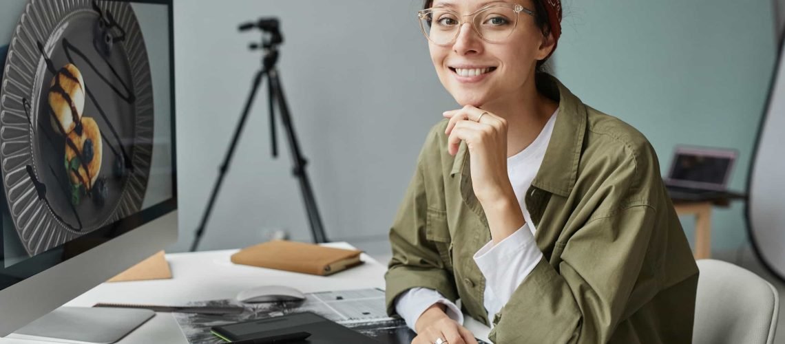 Person wearing glasses and a headband sits at a desk with a computer, smiling at the camera. Food appears on the screen, hinting at queens ny web design work. A tripod and laptop are visible in the background.