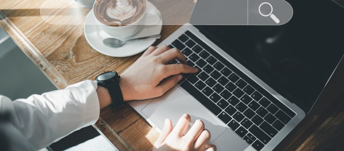 Person typing on a laptop at a wooden table with a cup of coffee, sunglasses, and plant nearby; a digital search bar graphic is overlaid on the image.