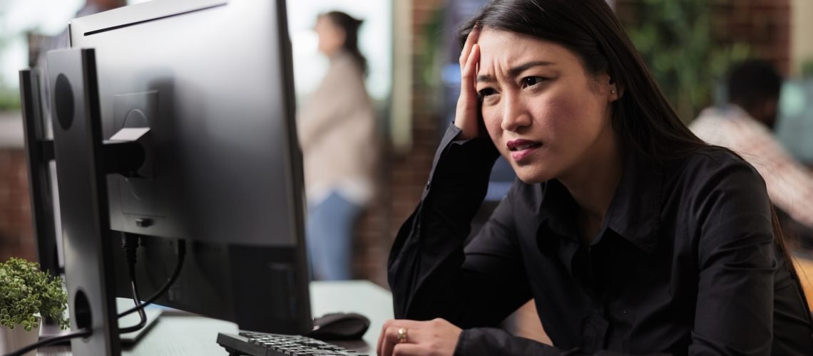 A woman sits at a desk in an office, looking frustrated while using a computer. Others are seen in the blurred background.