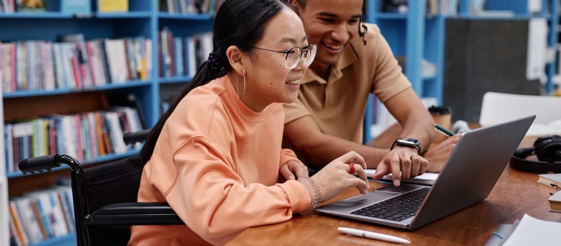 Two students, highlighting accessibility, work together on a laptop at a table in a library with bookshelves in the background; one student uses a wheelchair. WCAG
