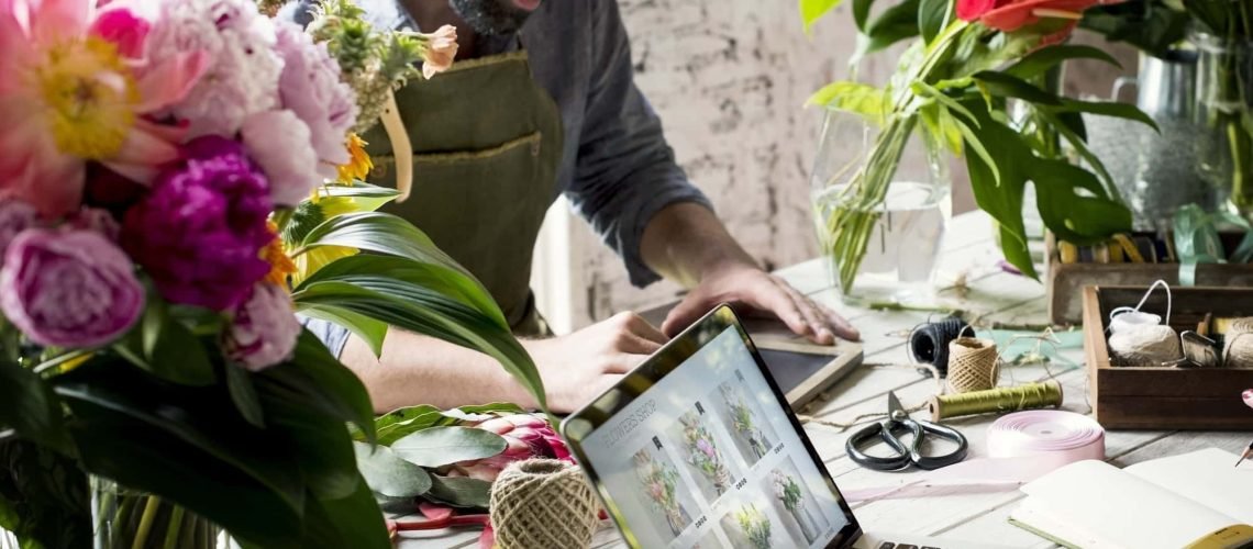 A florist in an apron arranges flowers at a table with a laptop and various floral tools in a bright workshop.