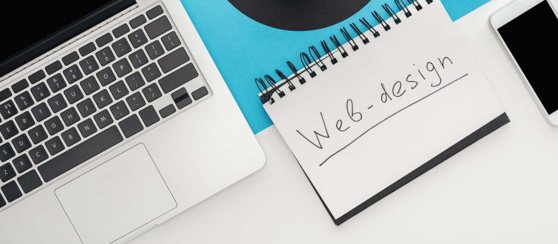 A laptop, smartphone, potted plant, and a spiral notebook labeled "Web-design" sit on a desk with a blue and white background—an inspiring NYC Web Designer workspace.