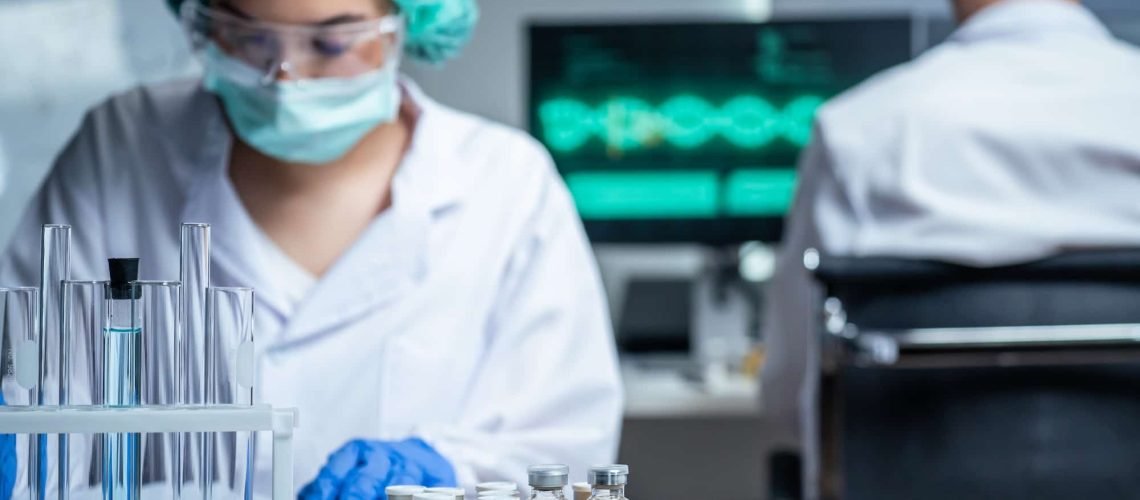 A scientist in protective gear works in a lab with test tubes, while several COVID-19 vaccine vials are displayed on the table in the foreground for a pharmaceutical website.