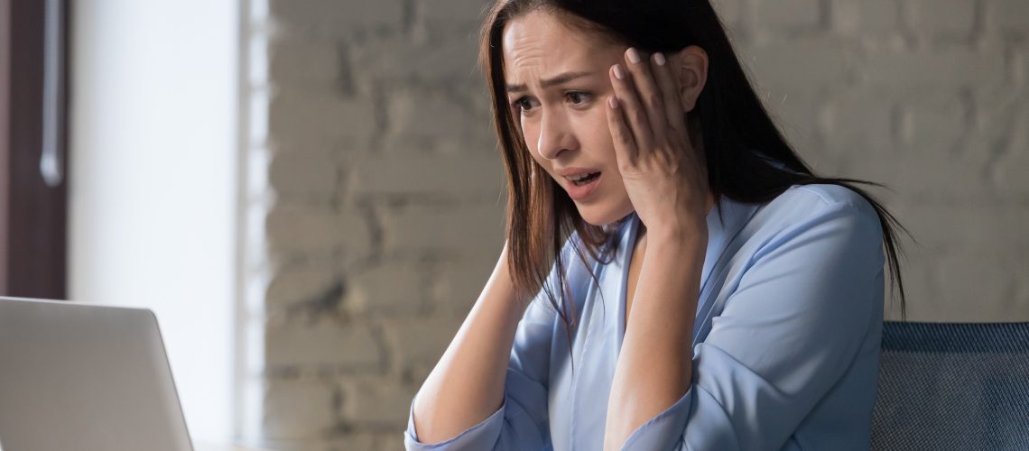A woman sits at a desk looking at a laptop with a worried expression, holding her head with both hands. The Hidden Costs of Using Wix and Squarespace for Your Business