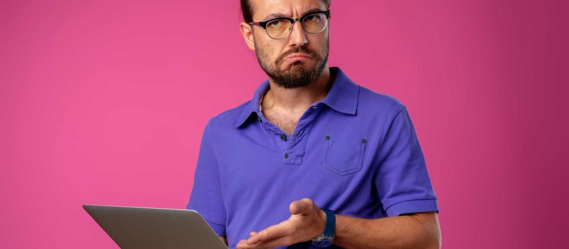 Man in a blue polo shirt and glasses holding a laptop, looking confused or frustrated, against a plain pink background.