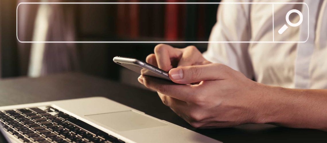 A person in a white shirt uses a smartphone next to an open laptop, with a digital search bar graphic overlay at the top of the image.