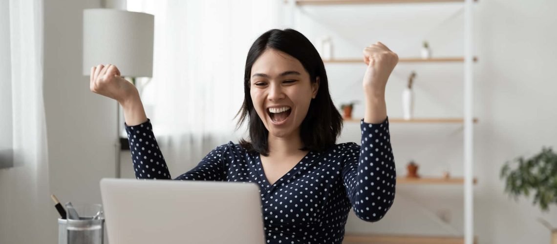 Person sitting at a desk with a laptop, raising fists in excitement.