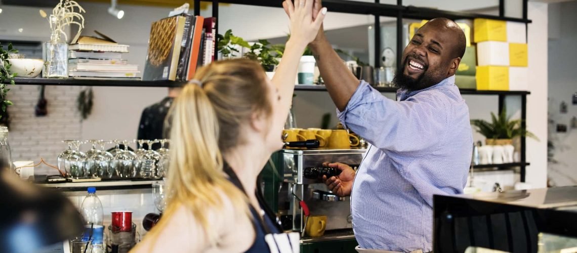 Two people wearing aprons share a high-five behind a coffee counter, smiling and celebrating their success in local SEO. Shelves with cups and books are in the background, reflecting their vibrant café's cozy charm.