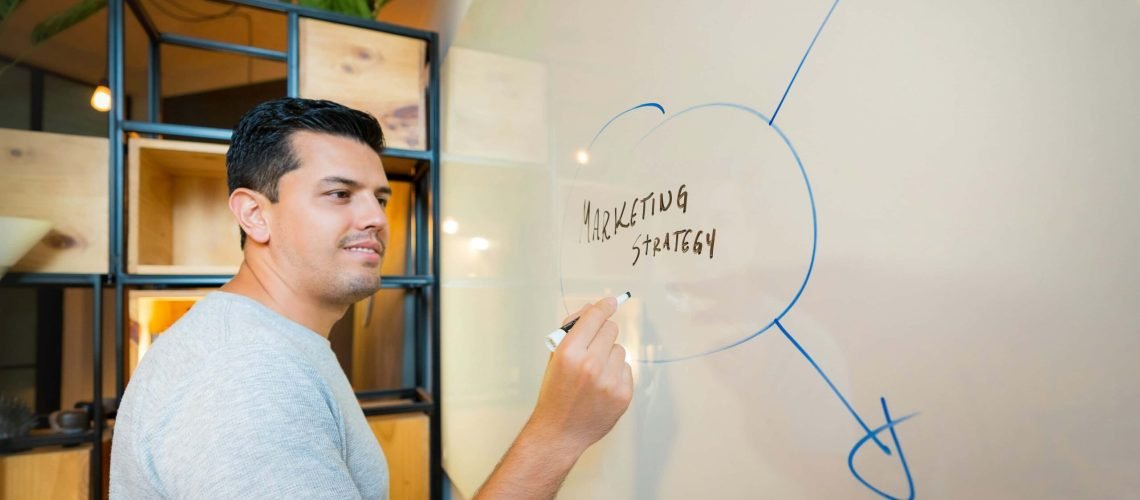 A person in a grey shirt is writing "Marketing Strategy" on a clear whiteboard with blue arrows extending from the text.