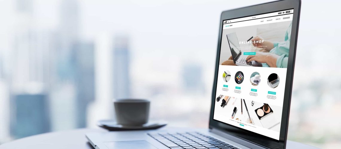 A laptop on a round table displays an online shop website; a cup and saucer sit nearby, with a blurred cityscape in the background.