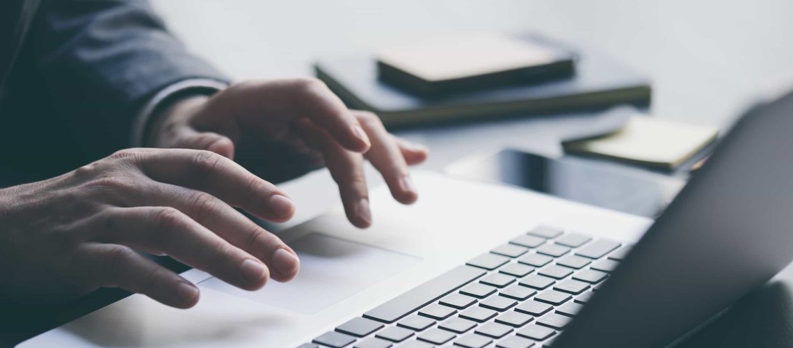 Close-up of a person’s hands typing on a laptop keyboard, with notebooks and a smartphone on the desk in the background.