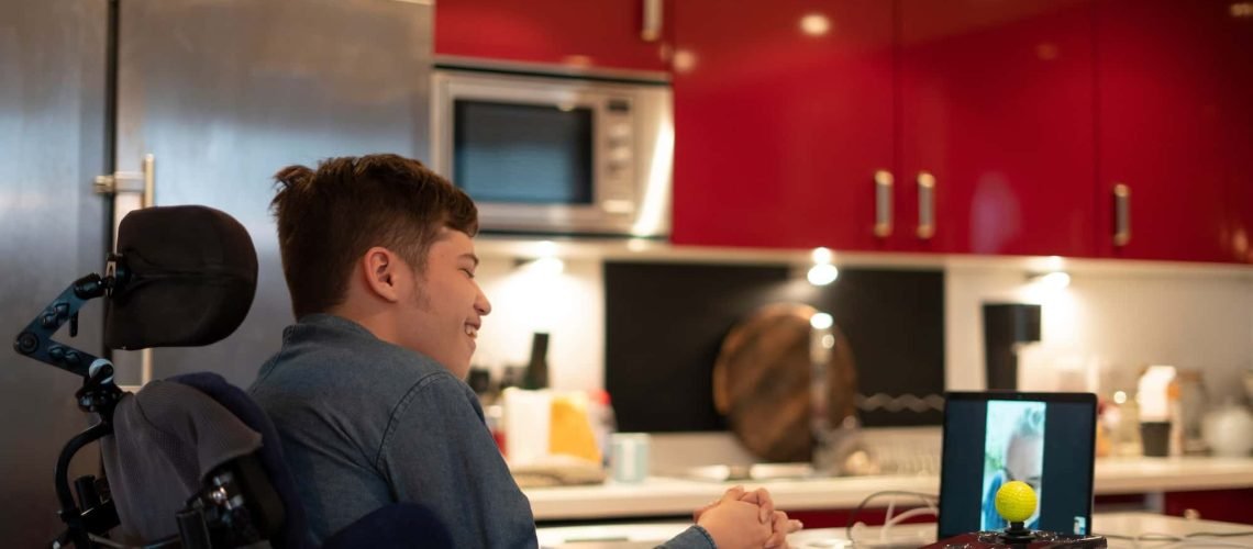 A person in a wheelchair smiles while facing a video call on a laptop in a modern kitchen with red cabinets.