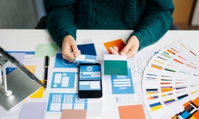 Person selecting color swatches at a desk with a smartphone, color samples, web design layouts, and a color palette fan spread out.