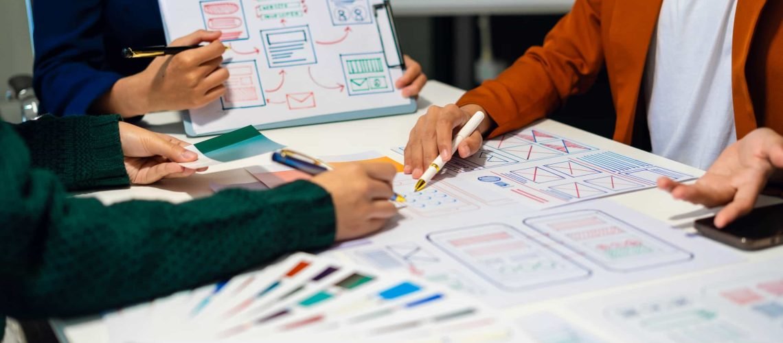 Three people work together at a table covered with papers, markers, and wireframe sketches, discussing and designing website or app layouts.