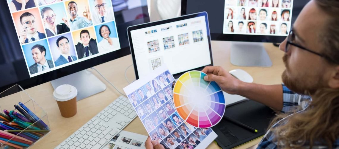 A person with long hair reviews a color palette and photos on multiple computer monitors, sitting at a desk with art supplies and a graphics tablet.